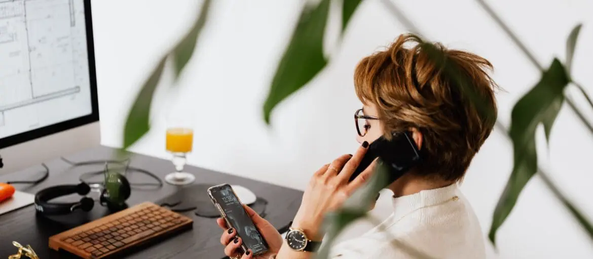 Woman looking at cellphone, while on the phone, in front of a computer